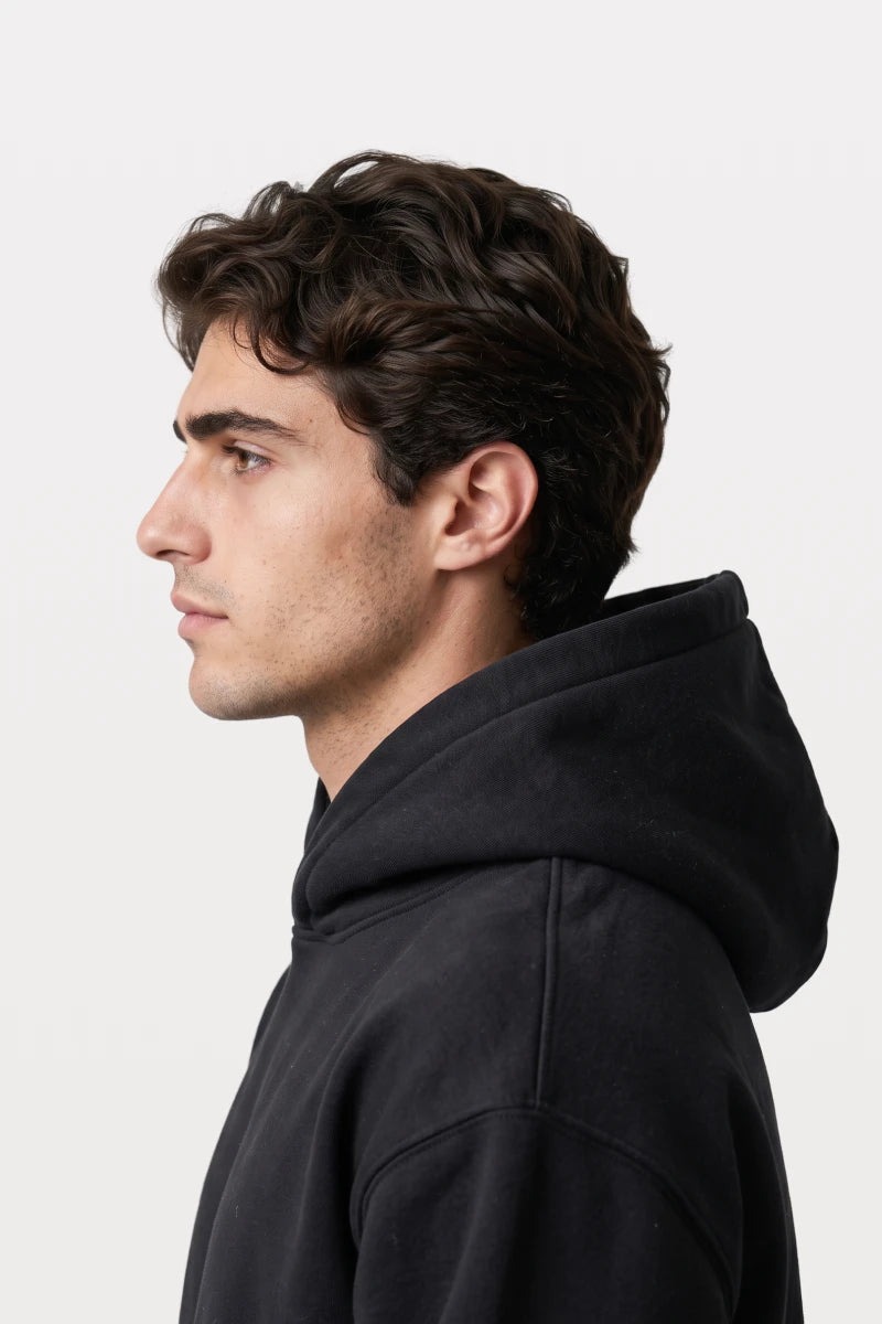 A man wearing a RAQI black essential pullover hoodie with a spacious hood, shown in a side profile view.