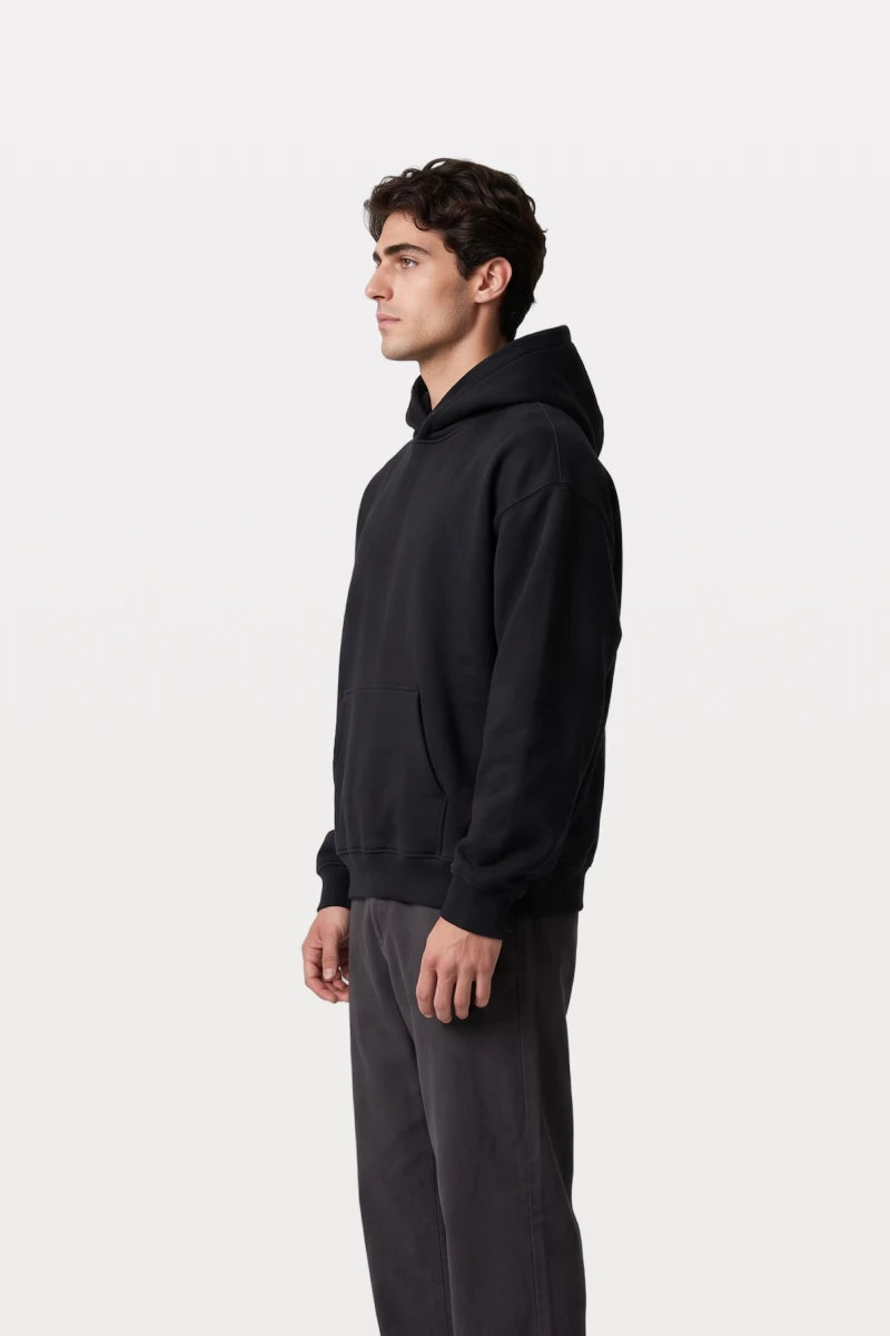 A man wearing a RAQI black essential pullover hoodie with a relaxed fit, hood, and kangaroo pocket, side profile view.