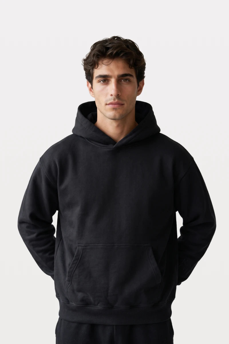 A man wearing a RAQI black essential pullover hoodie with a relaxed fit, kangaroo pocket, and ribbed hem.