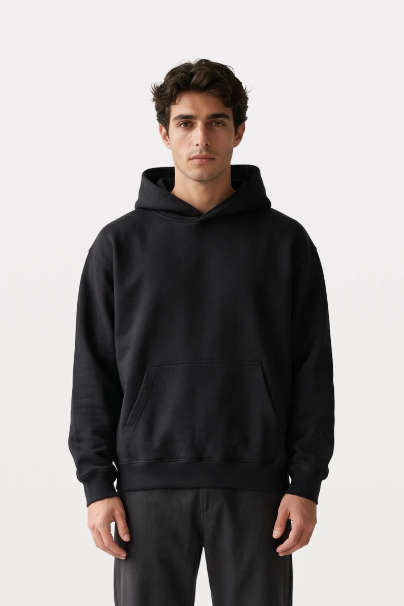 A man wearing a RAQI black premium cotton pullover hoodie with a relaxed fit, kangaroo pocket, and ribbed cuffs.