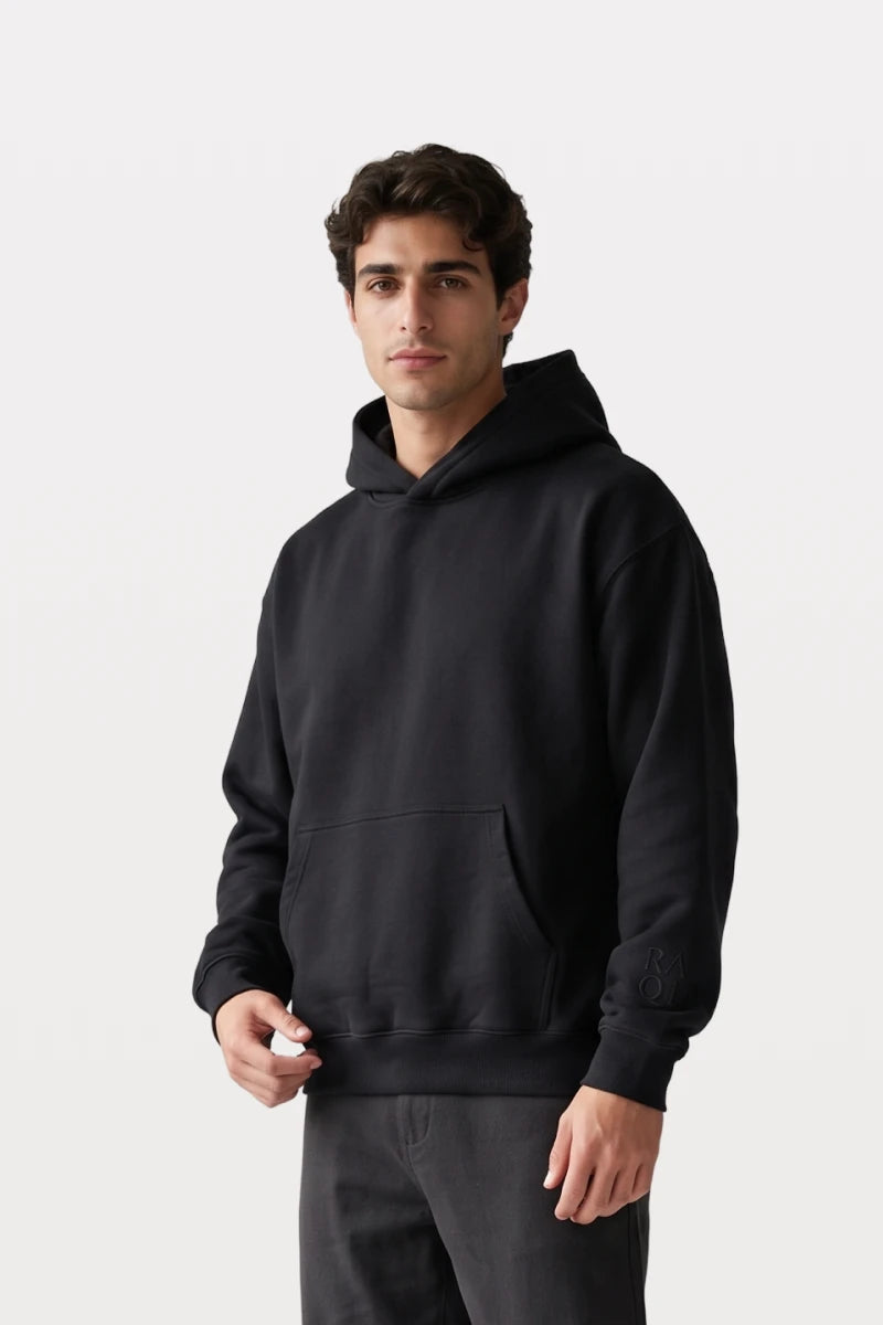 A man wearing a RAQI black relaxed-fit pullover hoodie with a kangaroo pocket and tonal sleeve branding.