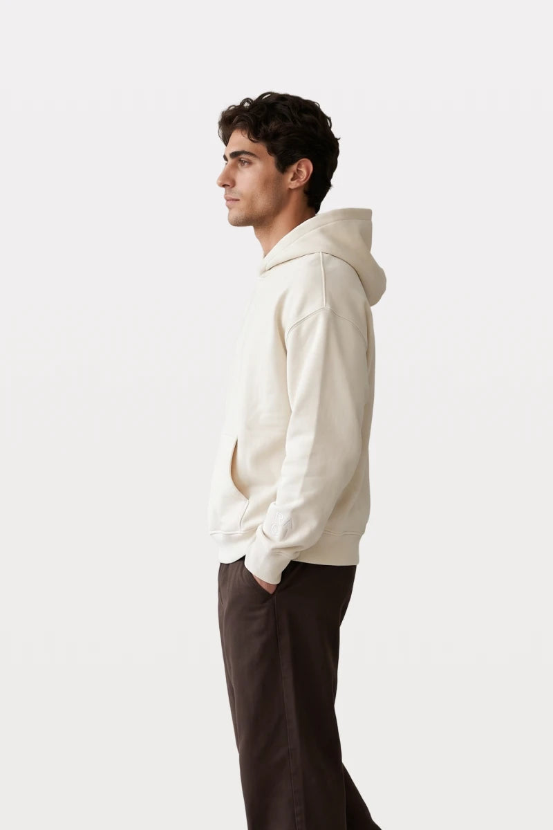 A man wearing a RAQI creamy light beige heavyweight pullover hoodie with a kangaroo pocket, side profile view.