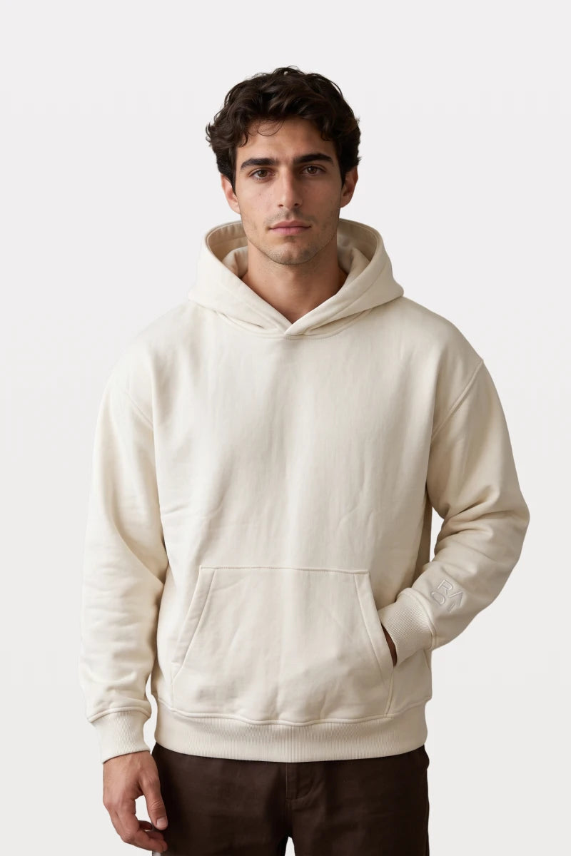 A man wearing a RAQI creamy light beige heavyweight pullover hoodie with a kangaroo pocket and ribbed cuffs.